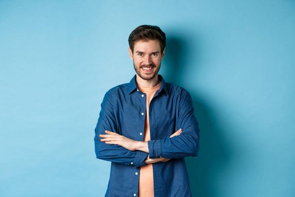 Selbstbewusster junger Mann, der sich wie ein Profi fühlt, die Arme auf der Brust verschränkt und frech lächelt, steht auf blauem Hintergrund. - Foto, Bild
