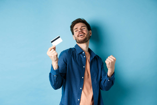 Happy and satisfied young man dancing with plastic credit card, fist pump and smile, standing on blue background - Fotografie, Obrázek