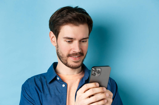 Close up portrait of happy young man reading mobile sreen, networking in smartphone, standing on blue background - Zdjęcie, obraz