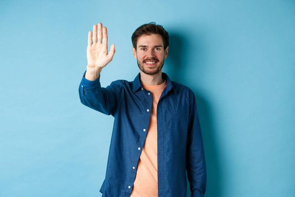 Freundlicher junger Mann lächelt und hebt die Hand für High Five, verzichtet auf Begrüßung oder Hallo, steht auf blauem Hintergrund - Foto, Bild