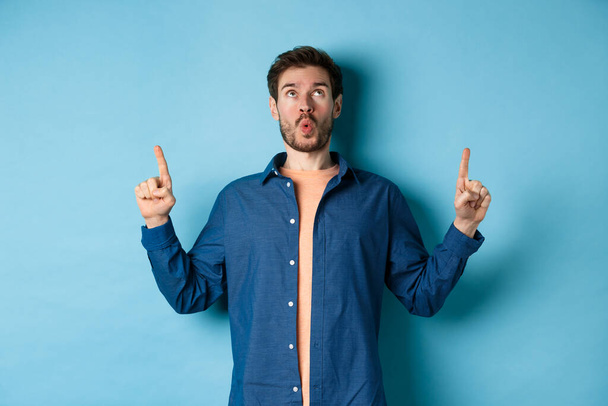 Image of surprised young man say wow, looking and pointing fingers up at promo offer, standing on blue background - Photo, Image