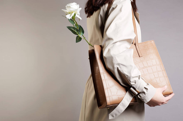 žena v módním plášti a tašce s bílou růžovou květinou. Módní dívka drží kabelku - Fotografie, Obrázek