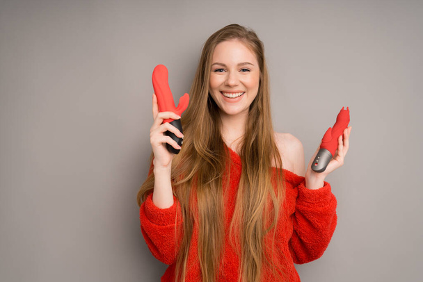 A girl of Caucasian appearance smiles broadly and holds two dildos or vibrators for sex on a gray background - Valokuva, kuva