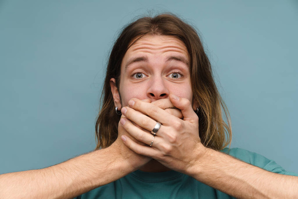Young white man wearing t-shirt covering his mouth and looking at camera isolated over blue wall - Foto, Bild