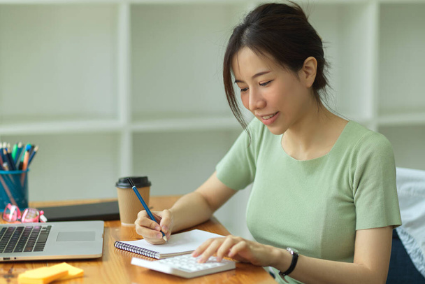 Kaunis nuori aasialainen naisyrittäjä, joka laskee kotitalouksien rahoituksen ja yrityksen lainan ja kirjoituslistan muistilehtiöissä nykyaikaisessa toimistotilassa - Valokuva, kuva