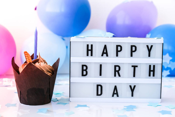 テキスト付きのライトボックスハッピー誕生日とチョコレートカップケーキキャンドル抽象的に焦点を当てた休日のためのぼやけたお祝いの背景。青い風船とコンフェッティ、誕生日ケーキ。グリーティングカードボーイコンセプト - 写真・画像