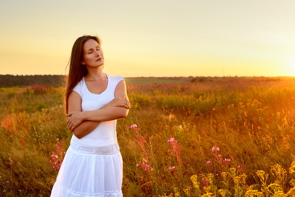 Jovem mulher de roupas brancas em pé no campo ao pôr do sol
 - Foto, Imagem