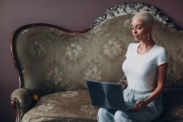 Mujer joven millenial con retrato corto de pelo rubio sentado con computadora portátil en un sofá vintage. - Foto, Imagen