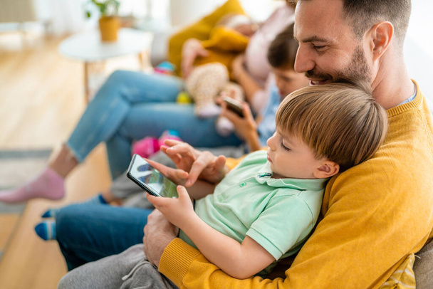Счастливые отец и сын играют в игры на мобильном телефоне дома. Технологическая зависимость - Фото, изображение