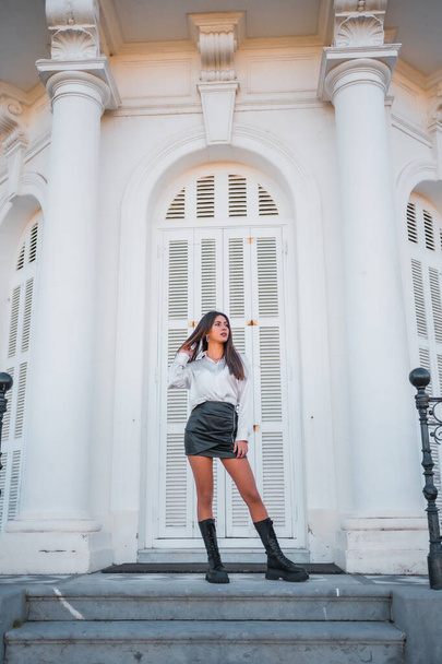 Lifestyle, a young brunette in a black leather skirt and shirt on the stairs of a white house - Фото, изображение