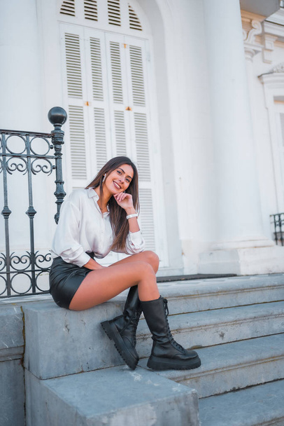 Lifestyle, fashionable pose of a young brunette in a black leather skirt and shirt smiling on some stairs one autumn afternoon, vertical photo - Photo, Image