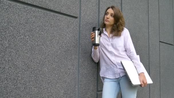 Gümüş dizüstü bilgisayarı ve kahve fincanı olan genç bir kadının portresi. Sabah koyu gri duvarın yanında toplantı bekliyor. Uzaktan çalışma, kahve molası, zeki öğrenci konsepti. - Video, Çekim