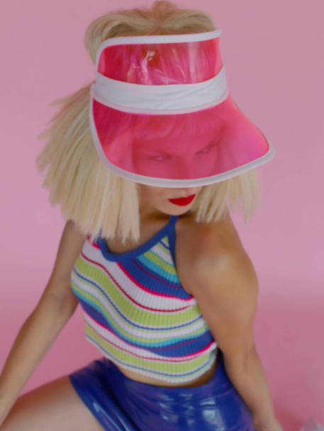 Uma menina na moda em um visor de tênis rosa transparente olha para a distância. Maquiagem brilhante com batom vermelho listrado top tanque: azul turquesa listra rosa. Cabelo loiro curvilíneo. - Foto, Imagem
