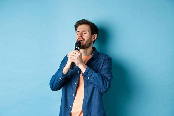 Romantik adam mikrofonda şarkı söylüyor, sahnede şarkı söylüyor, mavi arka planda duruyor. - Fotoğraf, Görsel