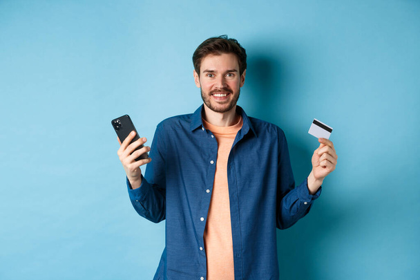 Koncepcja handlu elektronicznego. Podekscytowany młodzieniec ze smartfonem i plastikową kartą kredytową, zakupy online, stojący na niebieskim tle - Zdjęcie, obraz