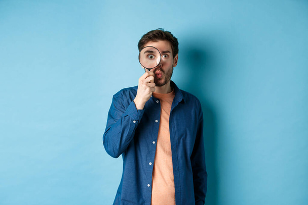 Image of surprised man found something interesting, say wow and look through magnifying glass, standing on blue background - Photo, Image