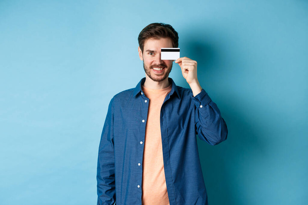 Felice cliente maschio in possesso di carta di credito in plastica su occhio e sorridente, in piedi in abito casual su sfondo blu - Foto, immagini