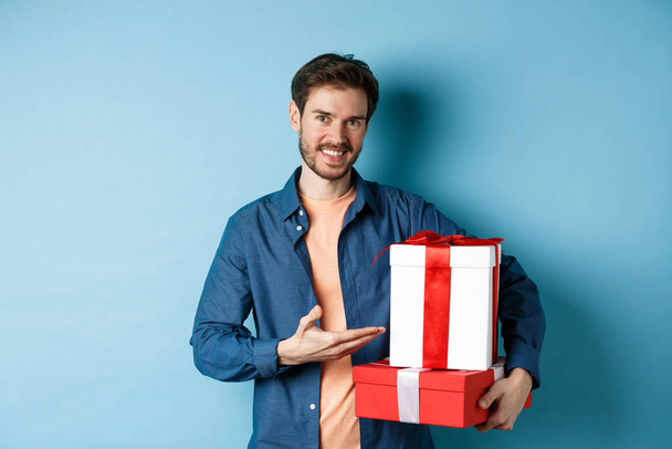 Young man in casual clothes buying romantic gifts on Valentines day, pointing at presents boxes and smiling, standing over blue background - Photo, Image
