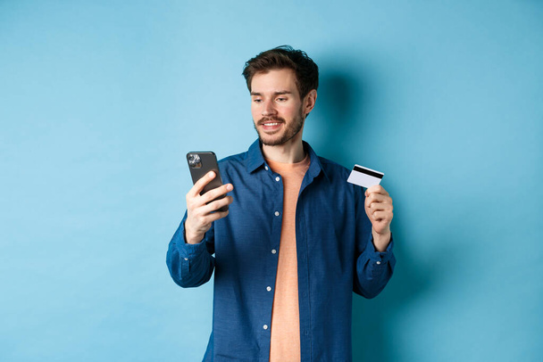 Концепция электронной коммерции. Изображение красивого кавказца, платящего онлайн, смотрящего на экран мобильного телефона и держащего пластиковую кредитную карту, синий фон - Фото, изображение