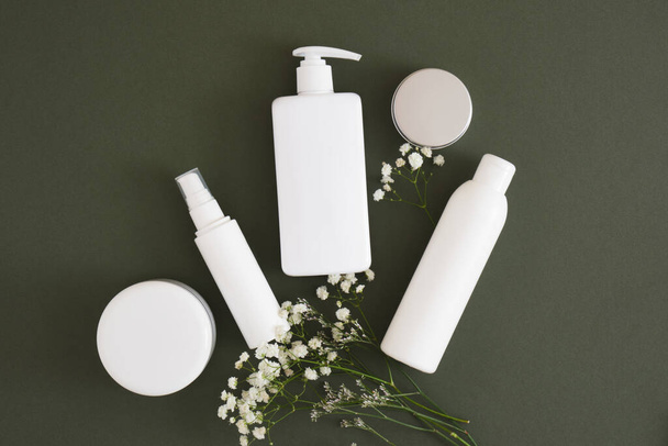 flowers, different cosmetic jars and three different blank white bottles on a dark green background backdrop Natural cosmetic packaging. mock up beauty product concept. - Photo, image