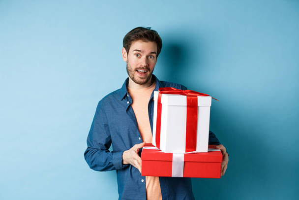 Schöner bärtiger Kerl mit Valentinstagsgeschenken für Liebhaber, der mit Geschenken in Schachteln steht und in die Kamera schaut, blauer Hintergrund - Foto, Bild