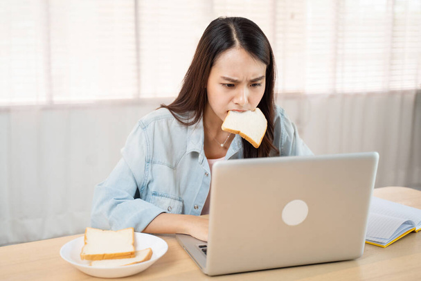 Asian attractive businesswoman hungry eating bread full mouth in hurry time while running business on notebook computer. Beautiful serious student feeling frustrated and busy working and study at home - Φωτογραφία, εικόνα