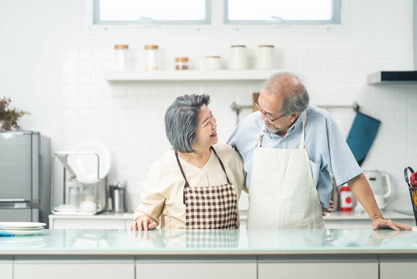 Ευτυχισμένο ζευγάρι ηλικιωμένων Ασιατών στην κουζίνα στο σπίτι απολαμβάνουν τη συνταξιοδότηση μαζί. Αγαπάτε τον παππού αγκαλιάζετε τη γιαγιά με ευτυχία και χαμόγελο, κοιτάζεστε. Σχέση δραστηριότητας στο σπίτι - Φωτογραφία, εικόνα