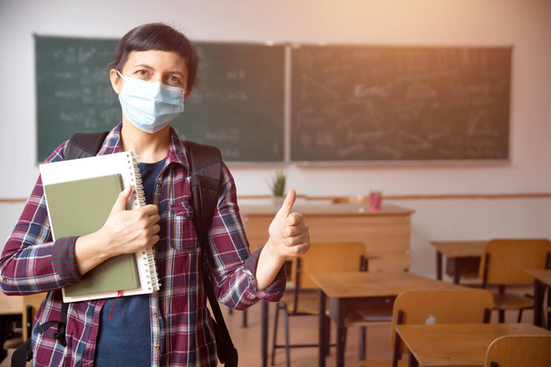 Smiling student girl wearing school backpack, mask and holding exercise book. Social distanting and classroom safety during coronavirus epidemic - Фото, изображение