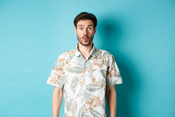 Summer holiday. Surprised tourist say wow and staring at camera, checking out awesome promo, standing in hawaiian shirt on blue background - Photo, Image