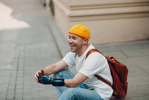 Jonge gehandicapte man met kunstmatige prothetische hand zitten op de stoep stad straat buiten en glimlachen. - Foto, afbeelding