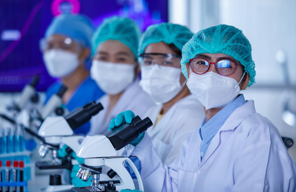 Skupina vědkyň nebo vědkyň s ochrannými hygienickými maskami a lékařskými uniformami, které spolu sedí a pracují s mikroskopy a zkumavkami v laboratoři a hledí do kamery. - Fotografie, Obrázek