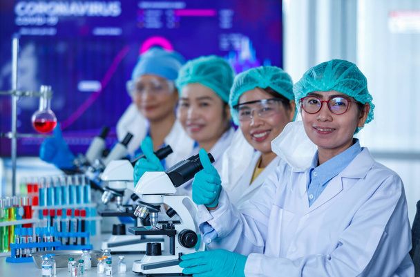 Gruppe von Forscherinnen oder Wissenschaftlerinnen mit Hygienemasken und medizinischen Uniformen sitzen zusammen und arbeiten im Labor mit Mikroskopen und Reagenzglas und blicken in die Kamera. - Foto, Bild