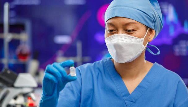 Женщины-медики в гигиенической маске и в покрытой одежде остаются в лаборатории, держат в руках и смотрят на бутылку вакцины Ковид-19 с лабораторными инструментами на заднем плане. Фокус на глазах. - Фото, изображение