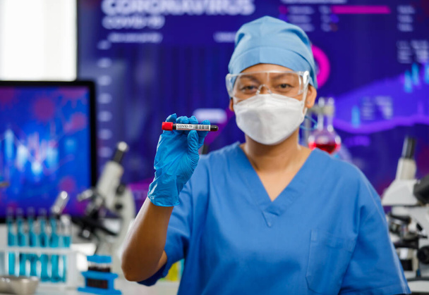 Vrouwelijke medische mensen met een hygiënisch gezichtsmasker en beschermend glas in de laboratoriumruimte, met monsterreageerbuis met laboratoriumapparatuur op de achtergrond en kijkend naar de camera. - Foto, afbeelding