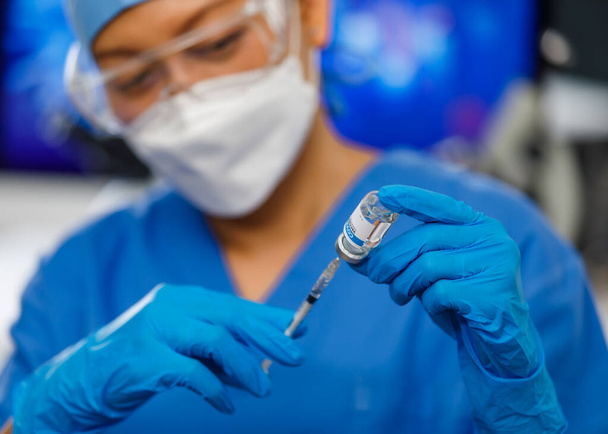 Gente médica en uniforme azul que usa mascarilla de higiene facial que sostiene el frasco de la vacuna e inserta la aguja en él, trabajando en laboratorio. - Foto, imagen