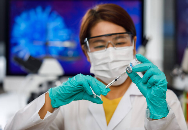 Médecins en uniforme blanc portant un masque d'hygiène du visage et des lunettes de protection tenant une bouteille de vaccin et y insérant une aiguille, travaillant en laboratoire. - Photo, image