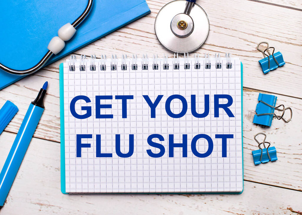 On a light wooden background, a stethoscope, a blue notepad, blue paper clips, a blue marker and a sheet of paper with the text GET YOOUR FLU SHOT. Medical concept - Foto, Imagem