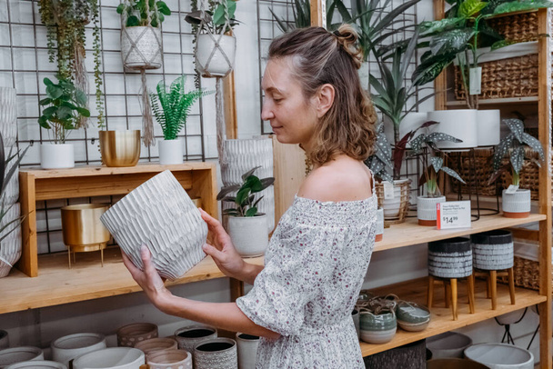 Ευχάριστη στιγμή για ψώνια σε offline κατάστημα. Νεαρή γυναίκα σε χαριτωμένο φόρεμα αγοράζει γλάστρα στο κατάστημα.Όμορφο κορίτσι επιλέγει φυσικό κομψό γλάστρες και φυτά για το διαμέρισμά της σε ένα μοντέρνο σπίτι έπιπλα - Φωτογραφία, εικόνα