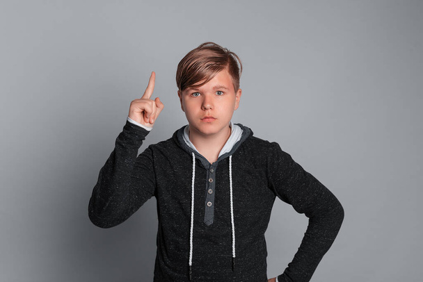 tiener jongen met ernstige gezichtsuitdrukking toont index vinger, het maken van aandacht gebaar, staan over grijze achtergrond, het dragen van casual kleding. Expressieve gezichtsuitdrukkingen - Foto, afbeelding