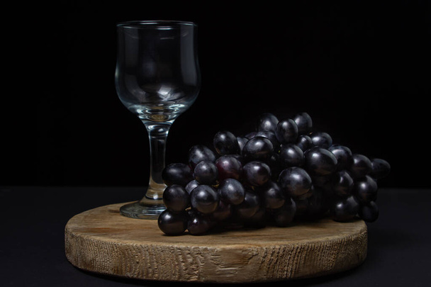 Czarne winogrona na czarnym tle. Grono winogron obok pustej szklanki na ciemnym tle - Zdjęcie, obraz