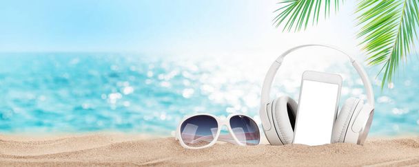 Smartphone, gafas de sol y auriculares en la playa tropical del mar con palmeras y arena brillante. Verano mar vacaciones, música y concepto de viaje. Con pantalla en blanco para tu app - Foto, imagen