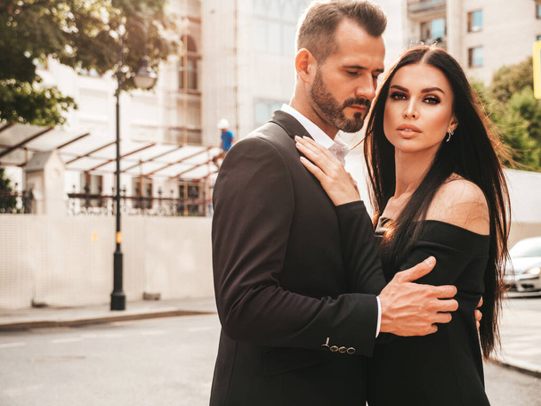 Beautiful fashion woman and her handsome elegant boyfriend in suit. Sexy brunette model in black evening dress. Fashionable couple posing in the street in Europe. Brutal man and his female outdoors - Zdjęcie, obraz
