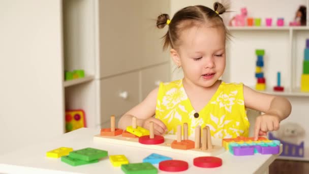 Uma menina sentada a uma mesa com vários brinquedos educativos de madeira está brincando com um novo popit de brinquedo de tendência moderna. A criança gosta de jogar pop-lo brinquedo e espinhas pop mais do que com pirâmides. Quarto de crianças - Filmagem, Vídeo
