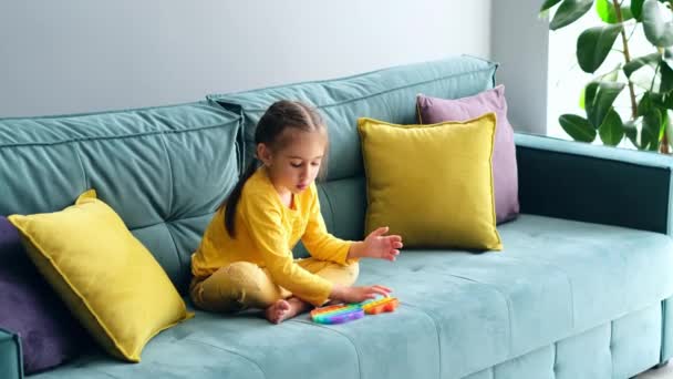 Ein Kind in gelben Freizeitklamotten sitzt auf dem Sofa und spielt mit dem beliebten Pop-It-Sinnesspielzeug. Spiele zur Entwicklung der Handmotorik. Grüne Couch mit gelben Kissen. Popitkunst - Filmmaterial, Video