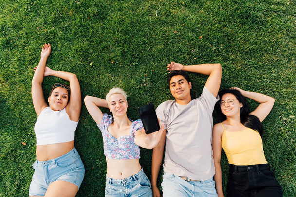 Gruppe junger multiethnischer Freunde im Freien, im Gras lächelnd, sich frei fühlend und entspannend beim Selfie mit dem Smartphone - Foto, Bild