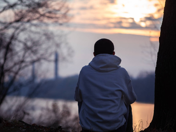 A lonely guy in a white hoodie is sitting on the river bank, cold autumn, sunset . Back view - Photo, Image