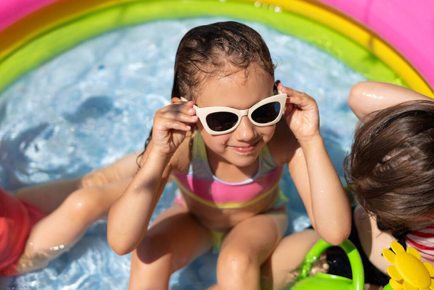 Мокра дівчина в сонцезахисних окулярах сидить у невеликому надувному басейні з чистою водою в гарячий сонячний день. Використовуючи захисний дитячий сонцезахисний крем. Діти розважаються у воді
. - Фото, зображення