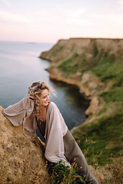 Stylish woman enjoys autumn nature at the edge of a rock above the sea, with blue sky and sea background. People, freedom, lifestyle, travel and vacations concept. - Photo, Image