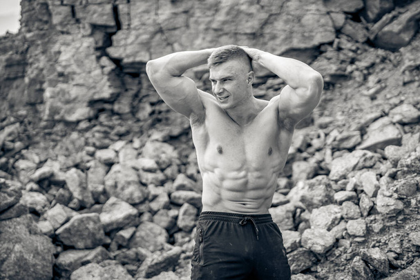 Bodybuilders pumping muscles outdoor. Man with perfect naked torso looking away while posing to the camera. Rocky background. Black and white photo - Foto, Imagen