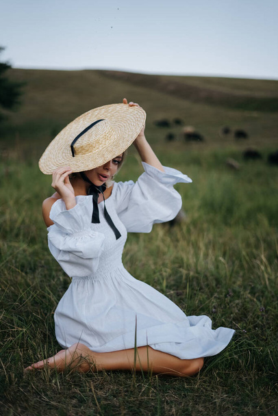молода красива модель дивиться на камеру. вона сидить на траві. літня концепція. жінка в білій сукні та солом'яному капелюсі
 - Фото, зображення